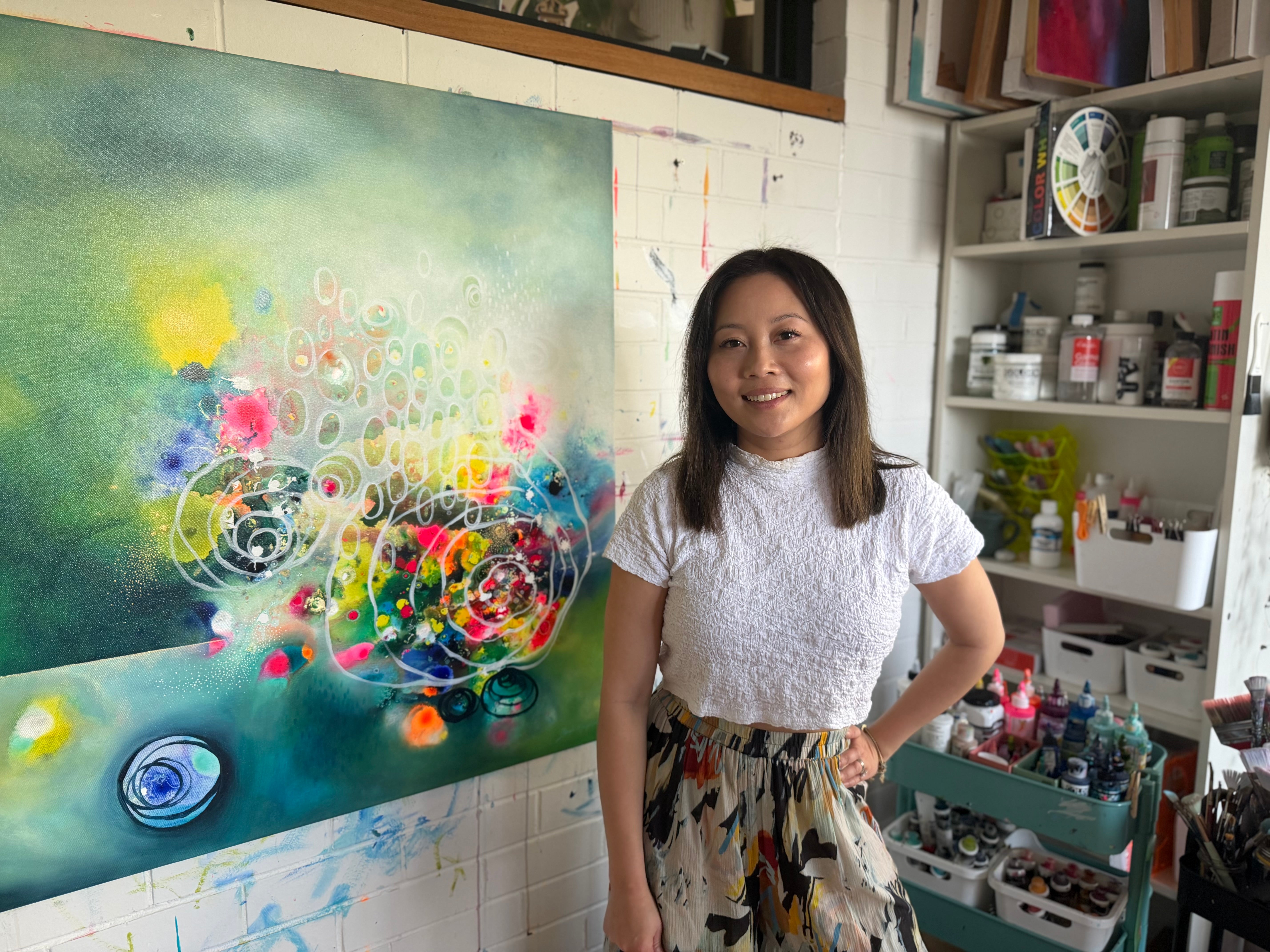 Brenda stands in her art studio next to a large colorful abstract painting, wearing a white textured short-sleeve top and a patterned skirt. Shelves filled with paint supplies are visible on the right.