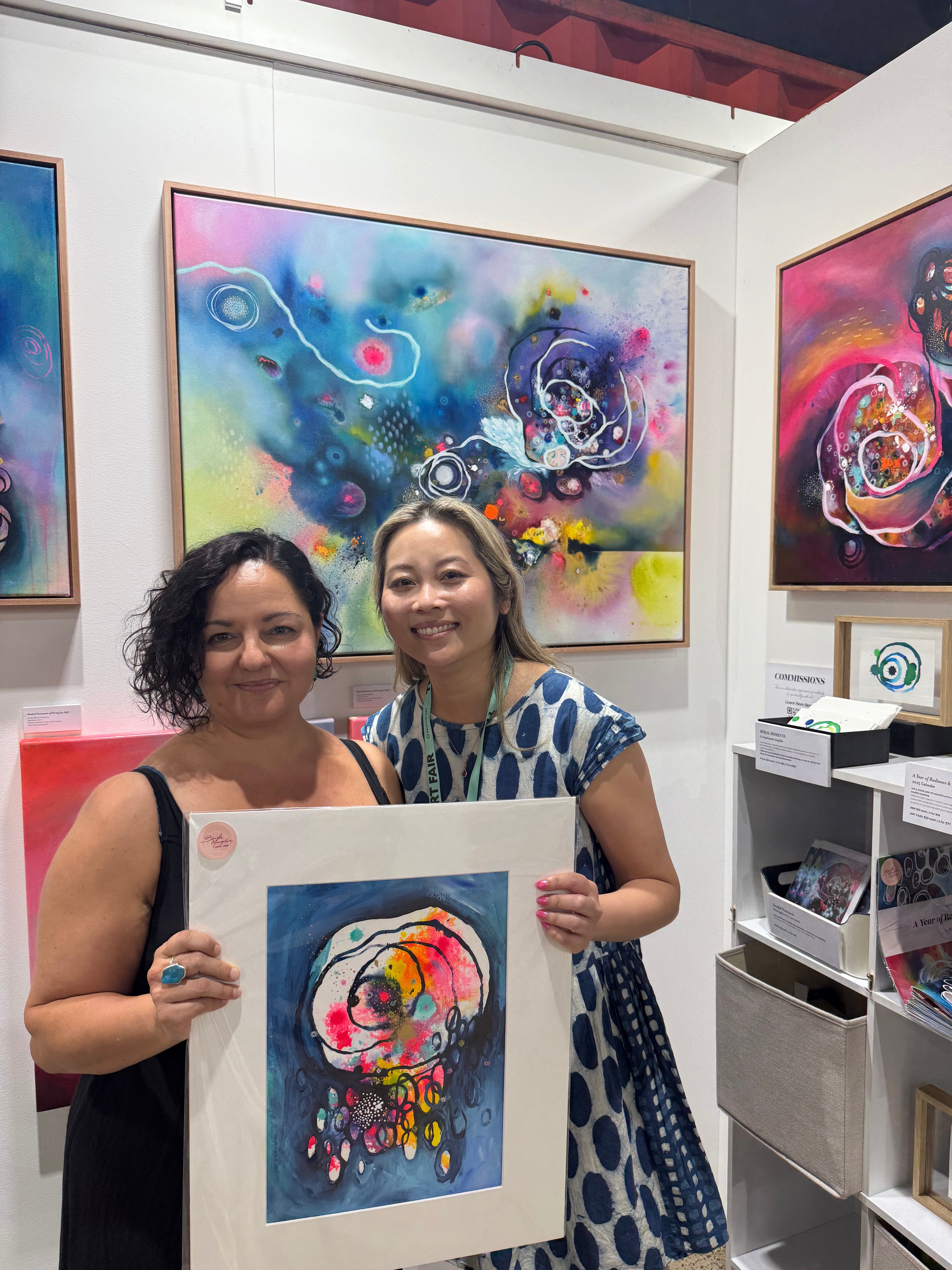 Two women standing in an art gallery booth, holding a colorful abstract painting print with a white mat. They are surrounded by other colorful abstract paintings on display